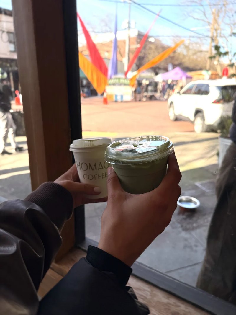 Holding an iced matcha and a coffee by a window at a street fair